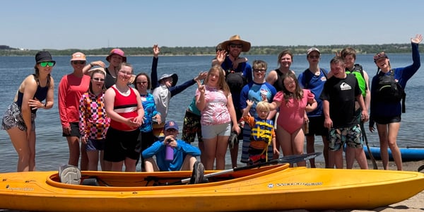 NSCD participants smiling and waving on a sandy lakeshore behind a yellow kayak during an NSCD group program.