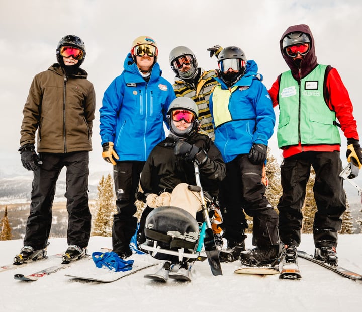 A skier using a sit-ski poses with five fellow skiers, including NSCD staff and a volunteer, on a snowy mountain slope.