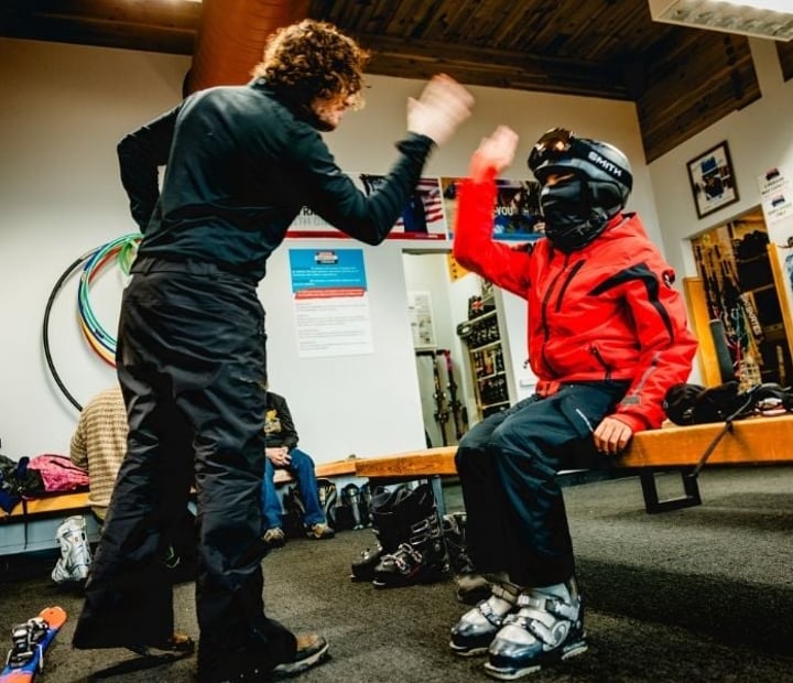 An NSCD participant and instructor share a high-five in the equipment room while preparing for an adaptive ski lesson.