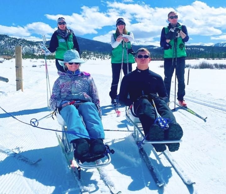 Two participants using adaptive Nordic sit-skis and three NSCD volunteers pose together on a groomed snow trail with mountain views in the background.