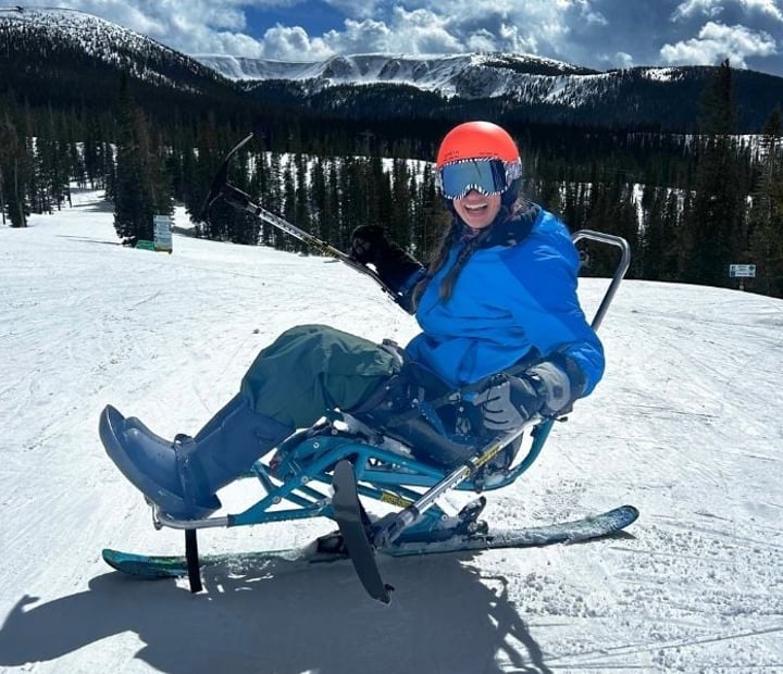 Participant using a sit-ski on a snowy ski slope with snow-covered mountains and evergreen trees in the background.