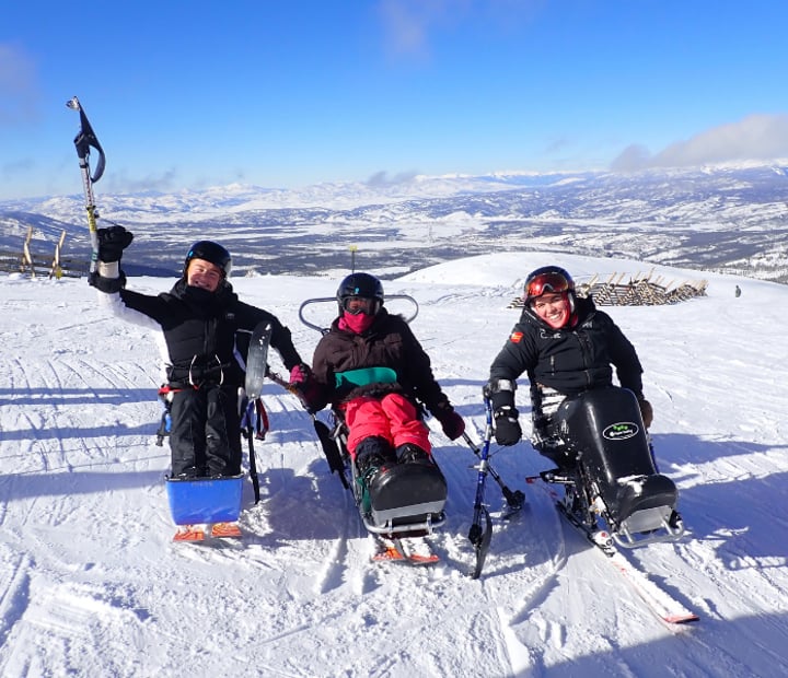 Three sit-skiers at the top of a mountain run raising their outriggers up in a happy pose.