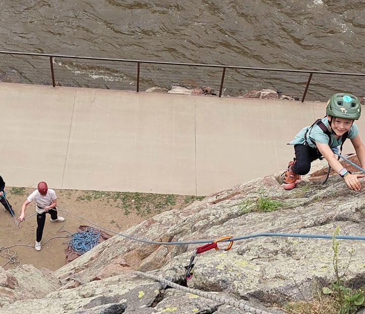 Young girl smiling and rock climbing with two belayers, a walkway, and a river below her.