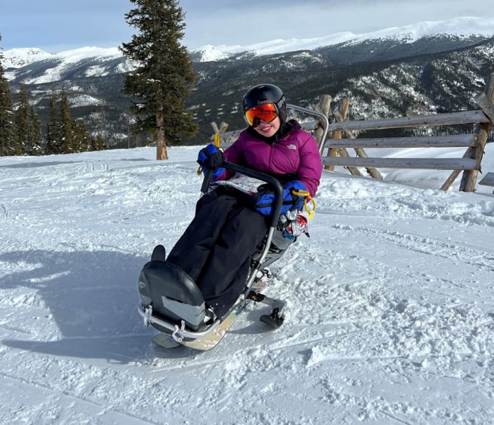 Female sit skier at the top of a run waiving to the camera.