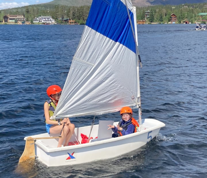 Two participants in a small white boat with blue and white sails, sailing on grand lake.