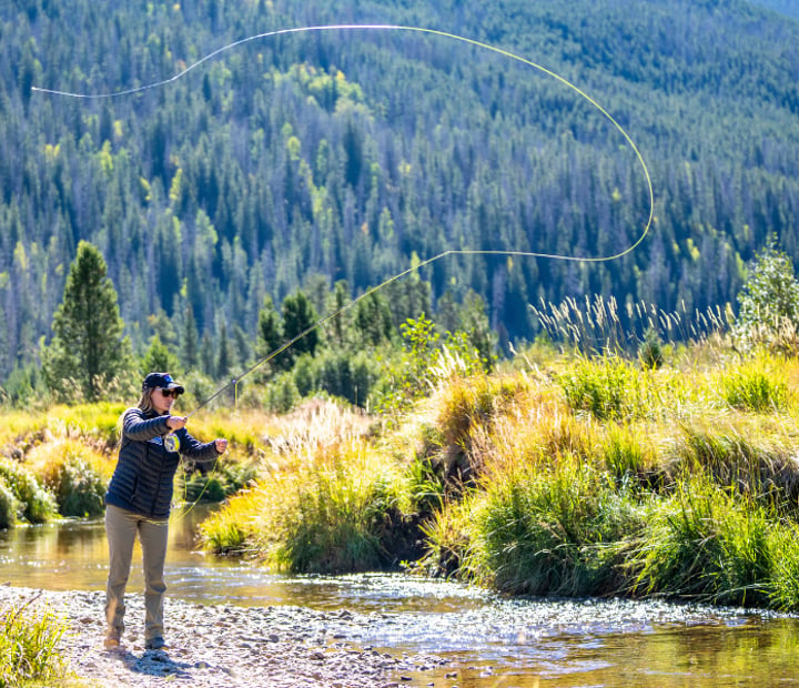Woman standing on the shore of a river, casting the line of a fly fishing rod with mountain hills in the background.