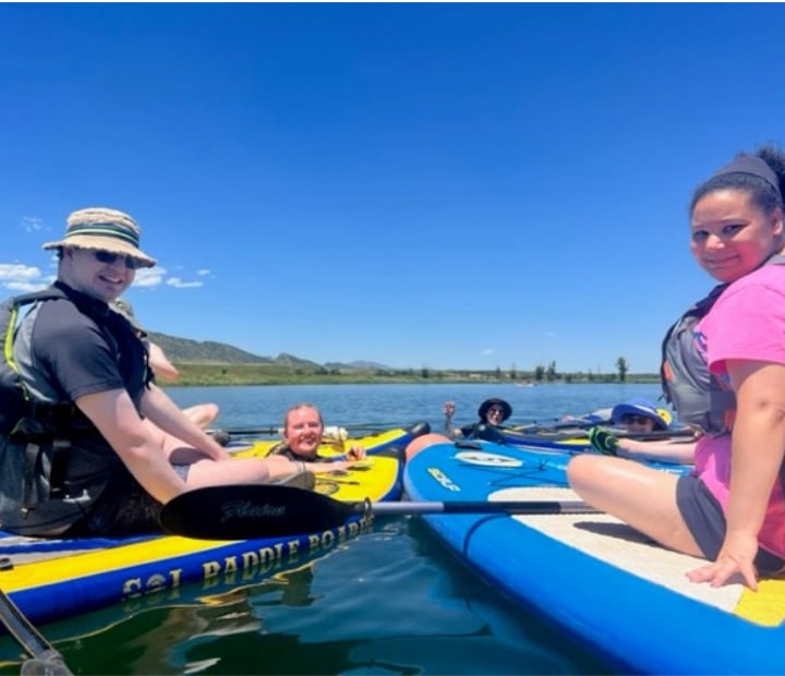 Two participants sitting on a paddleboard with three other participants swimming in the water near them.