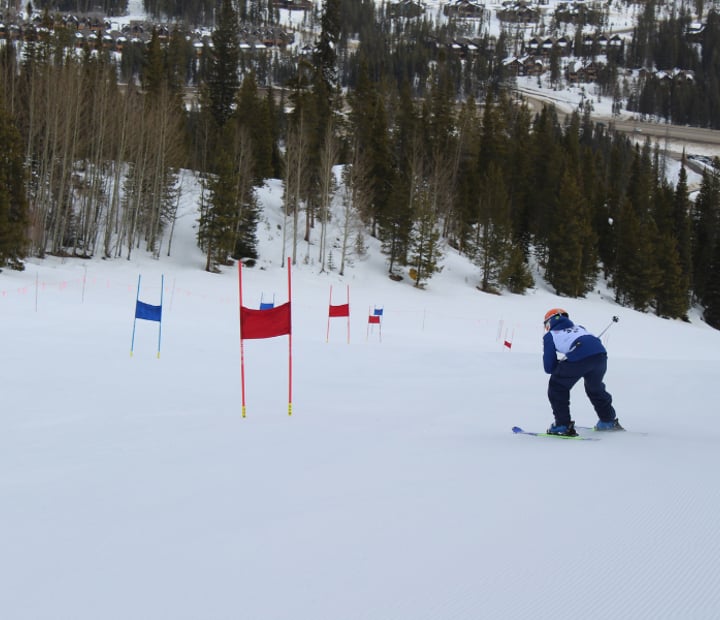 Ski participant in a tuck racing towards a red gate during race camp.