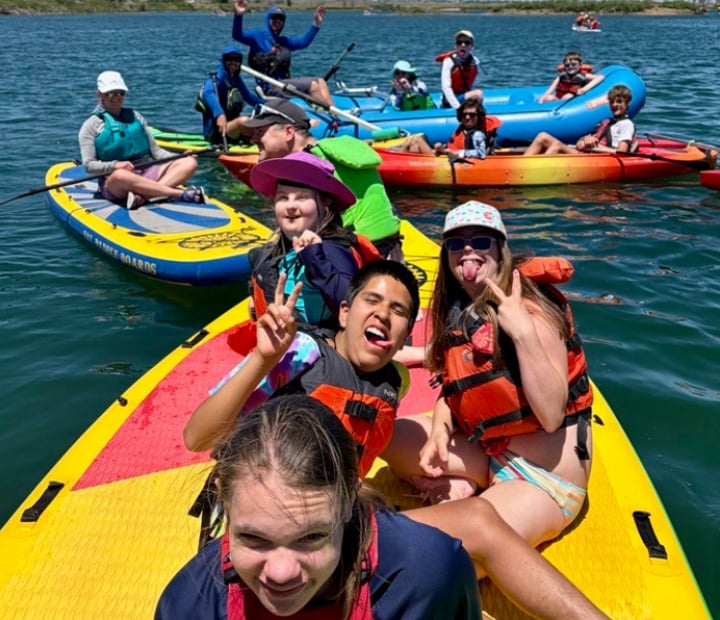 A group of youth participants wearing life jackets sit together on a large paddleboard during a water recreation program. A graphic on the image notes that the activity is for ages 8 and older.