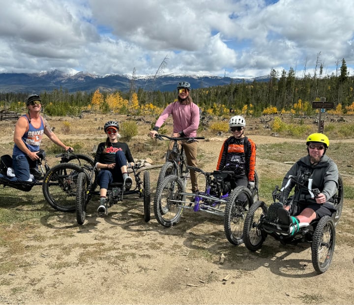 Adult participants gather with adaptive mountain bikes against a mountain backdrop during an adaptive bike program. The image indicates the activity is for ages 18 and older.