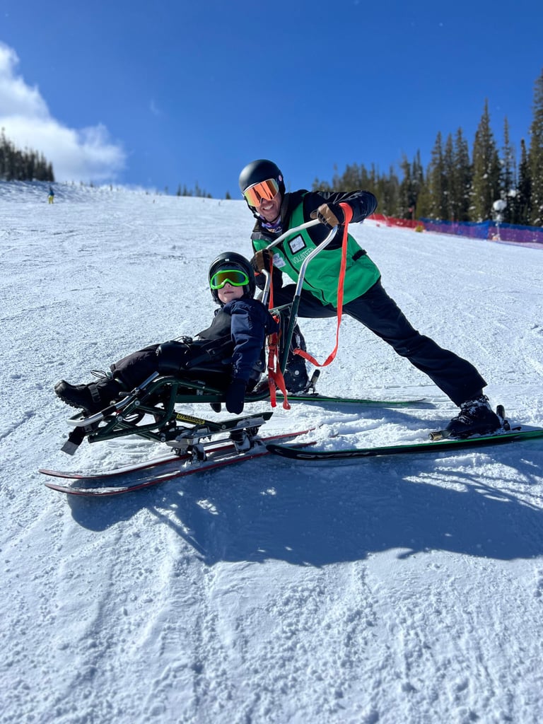 An NSCD volunteer uses a tether system to guide a participant in a sit-ski on a snow-covered ski run under a clear blue sky.