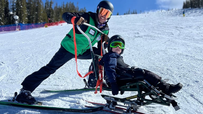 An NSCD participant rides a sit ski with the support of a volunteer instructor using guide handles on a snowy ski slope. Both wear helmets and goggles as they smile during an adaptive skiing lesson at Winter Park Resort.