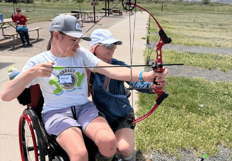 Dorian practices adaptive archery from his wheelchair with his mom, Kelli, supporting him during an outdoor NSCD program.