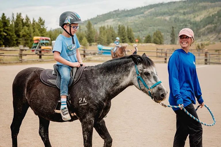 Child participant with a helmet riding a horse being steered by a smiling NSCD volunteer.
