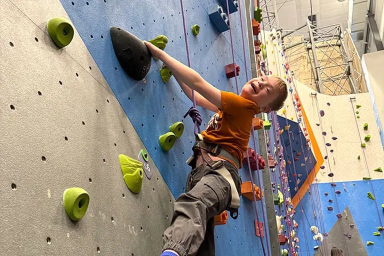 Child participant with rock climbing harness smiling while climbing up indoor climbing wall.