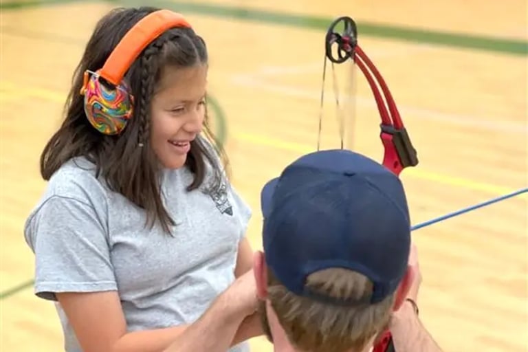 NSCD volunteer helping program participant to nock an arrow onto her bow in an indoor gym.
