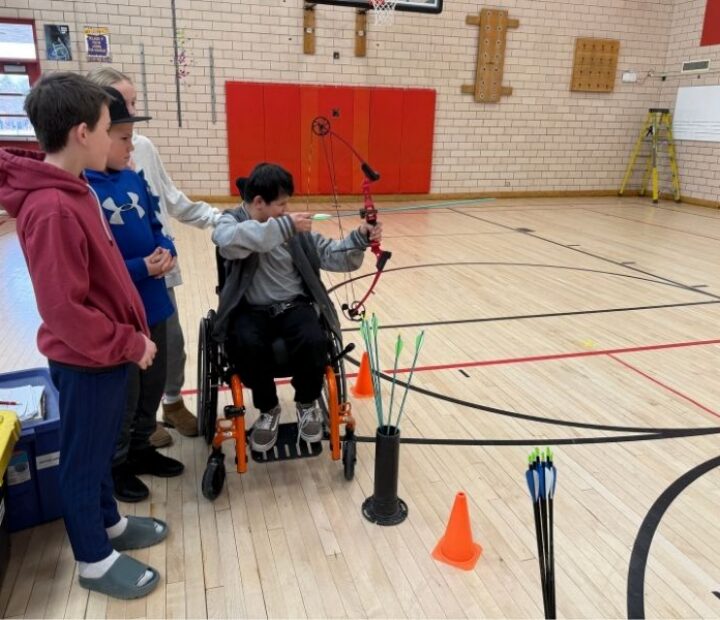 Participant in a wheelchair practices archery in a gymnasium while peers observe and offer support.