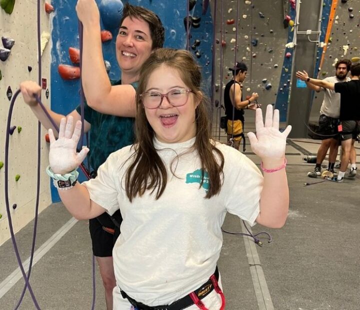 Participant wearing a climbing harness smiles and raises chalk-covered hands in an indoor climbing gym.