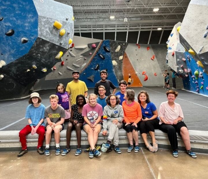 A group of participants and instructors pose together inside an indoor climbing gym with large bouldering walls in the background.