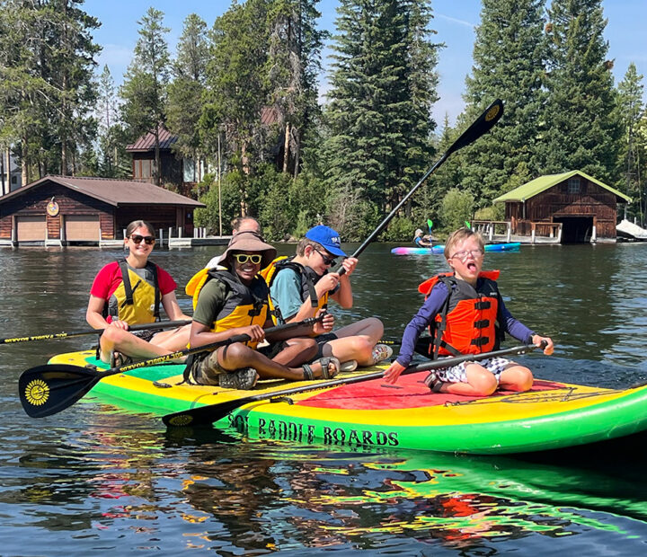 NSCD volunteer and participants on smiling large paddle board that is on a lake with pine trees in the background.