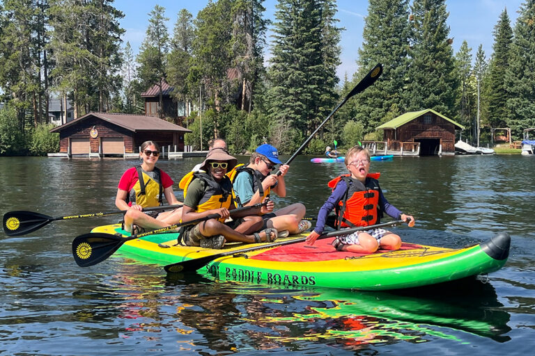 NSCD volunteer and participants on smiling large paddle board that is on a lake with pine trees in the background.