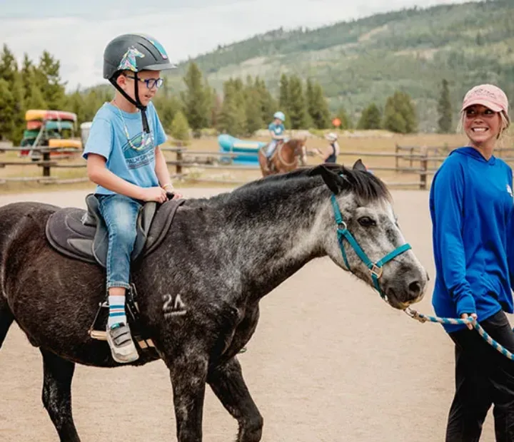 Child participant with a helmet riding a horse being steered by a smiling NSCD volunteer.
