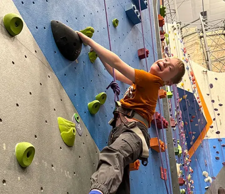 Child participant with rock climbing harness smiling while climbing up indoor climbing wall.