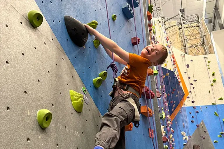 Child participant with rock climbing harness smiling while climbing up indoor climbing wall.