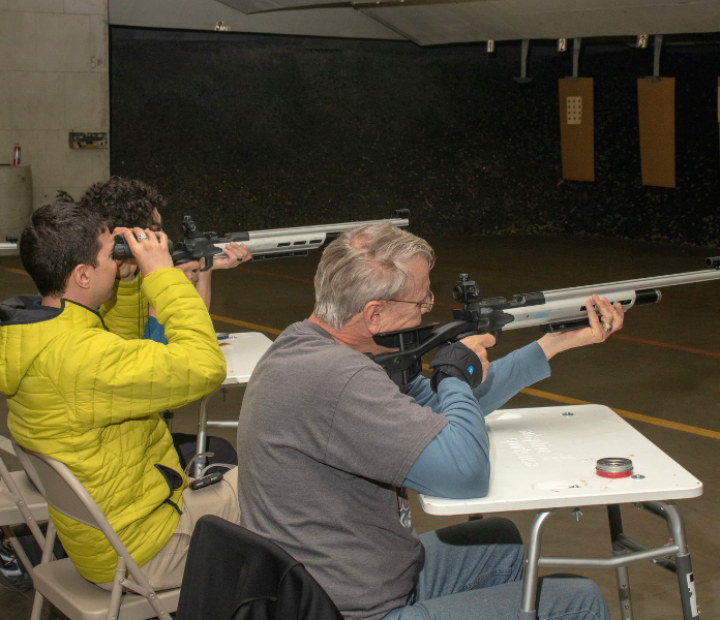 Group of men sitting at separate tables, aiming their air riffle guns towards their target, preparing to shoot.