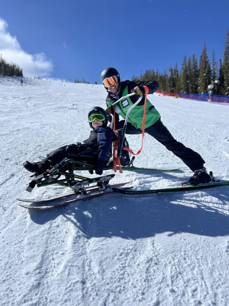 An NSCD volunteer uses a tether system to guide a participant in a sit-ski on a snow-covered ski run under a clear blue sky.