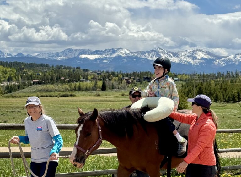 An NSCD participant smiles while riding a horse with adaptive support during an equine lesson. Two volunteers walk alongside the horse, one leading and one providing side support.