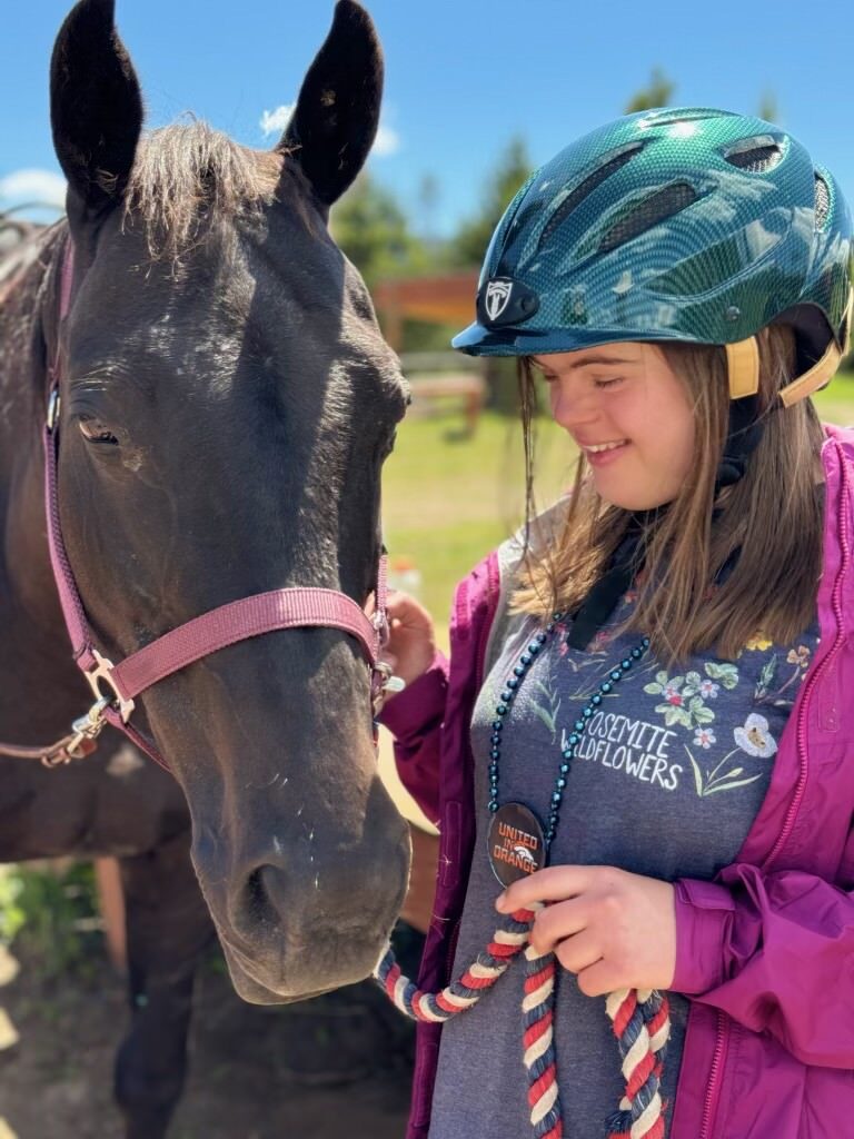 An NSCD participant wearing a riding helmet smiles while standing beside a horse during an adaptive horseback riding program.